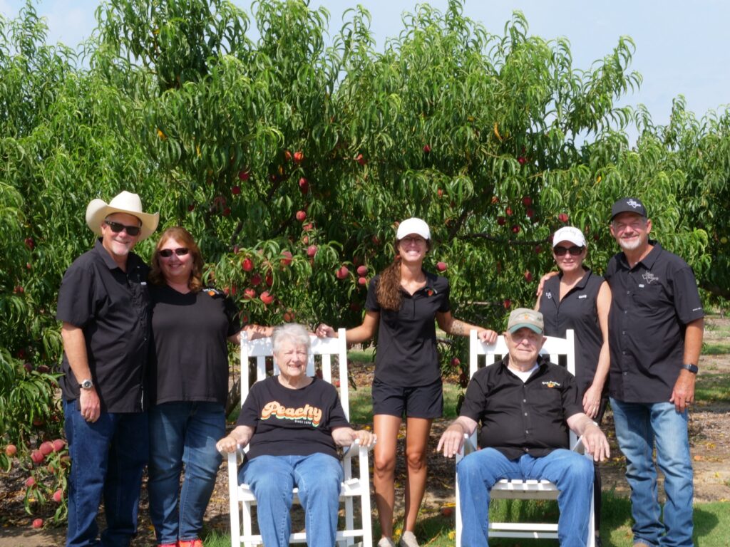 Picture of the Ham family behind Ham Orchard. Features founder Dale Ham and his wife Judy Ham and family with their daughters and extended family. Also, shows peach orchard in bloom in the background. Ham Orchards is located in Terrell, Texas and open from mid May to mid August for public visits.  